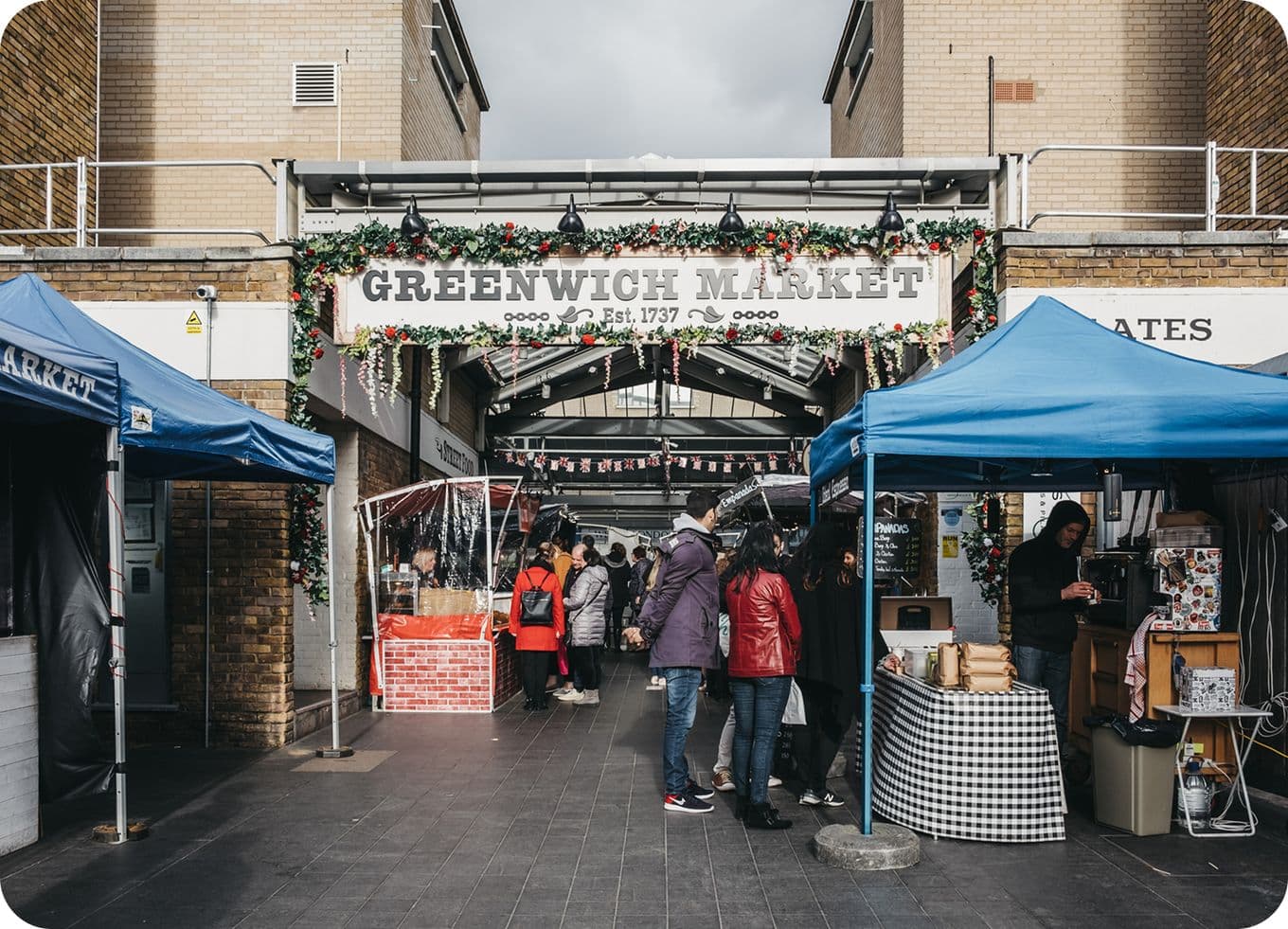 Greenwich Market entrance decorated with garlands, rows of stalls under blue canopies and shoppers browsing.