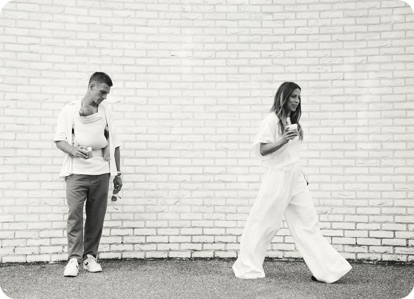A person in casual attire stands against a white brick wall, while another walks by holding a drink. Both are dressed in light-colored clothing.