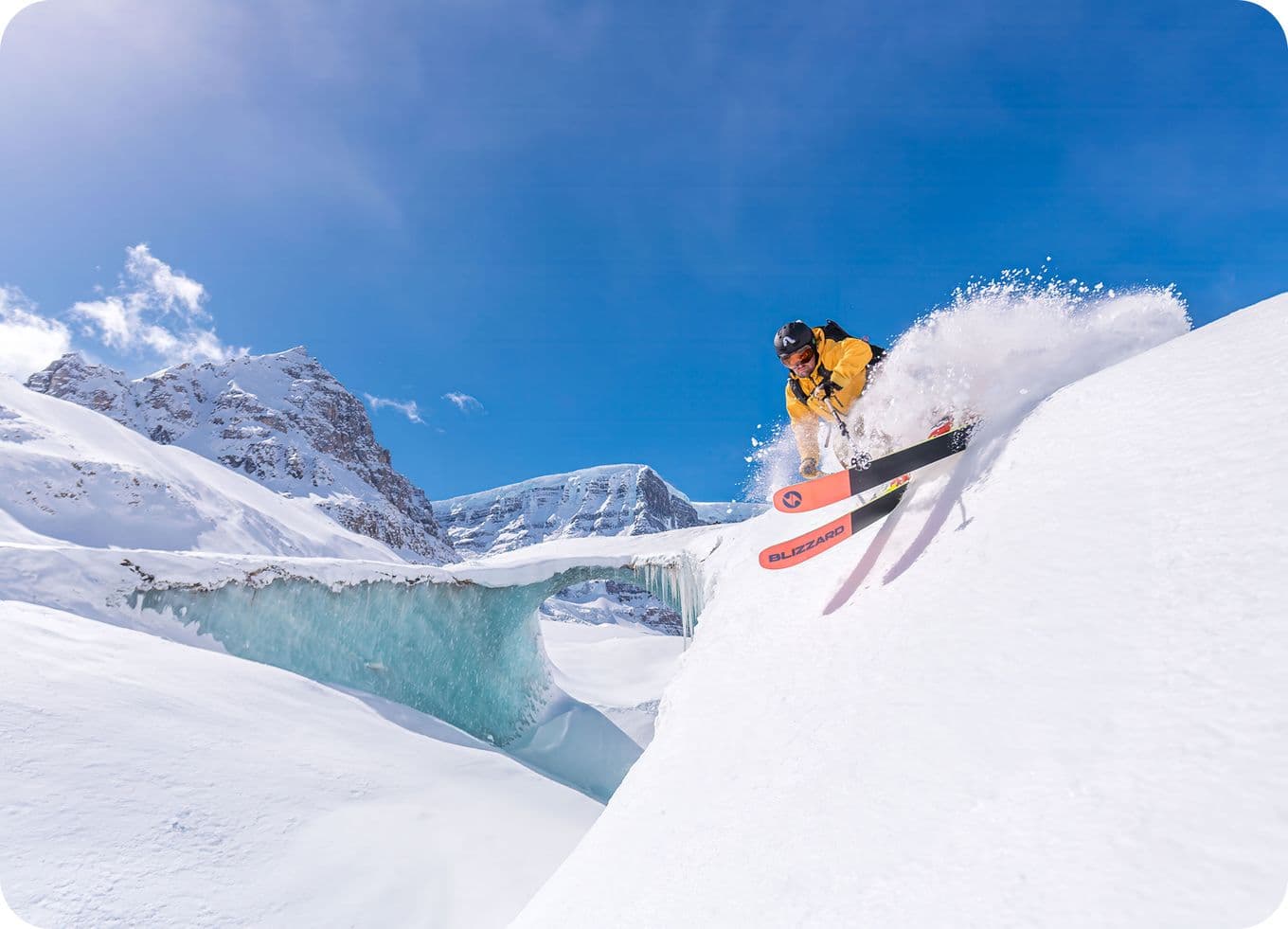 Skier in a yellow jacket descends a snowy slope, creating a spray of snow, with a backdrop of mountains and a clear blue sky.