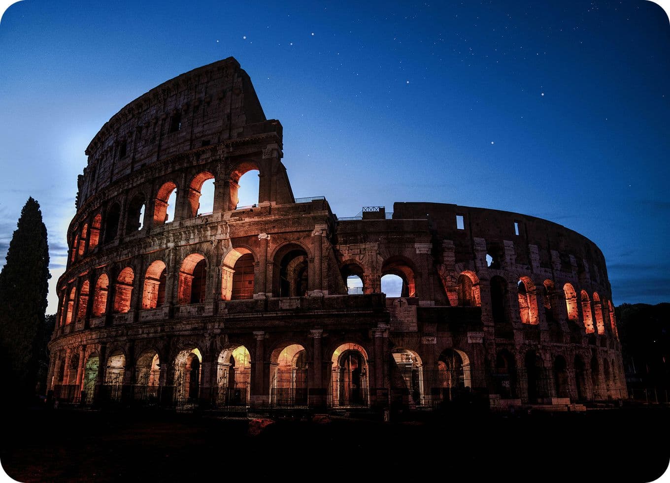 The Colosseum at night, illuminated with warm lights against a deep blue starry sky, showcasing its ancient architecture.