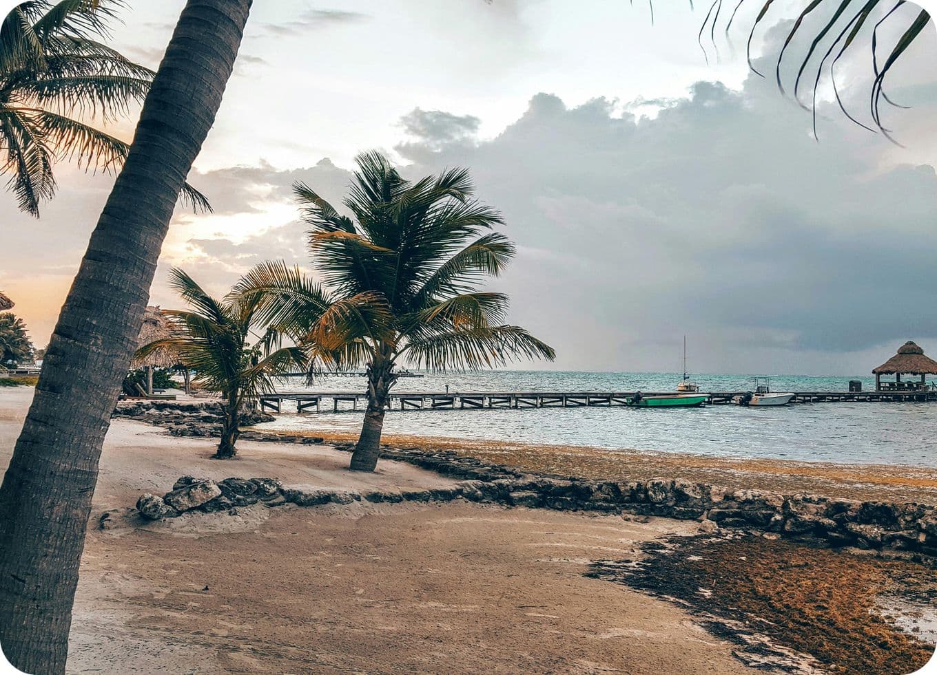 Tropical beach scene with palm trees, a wooden pier, and a small boat on the water under a cloudy sky at sunset.