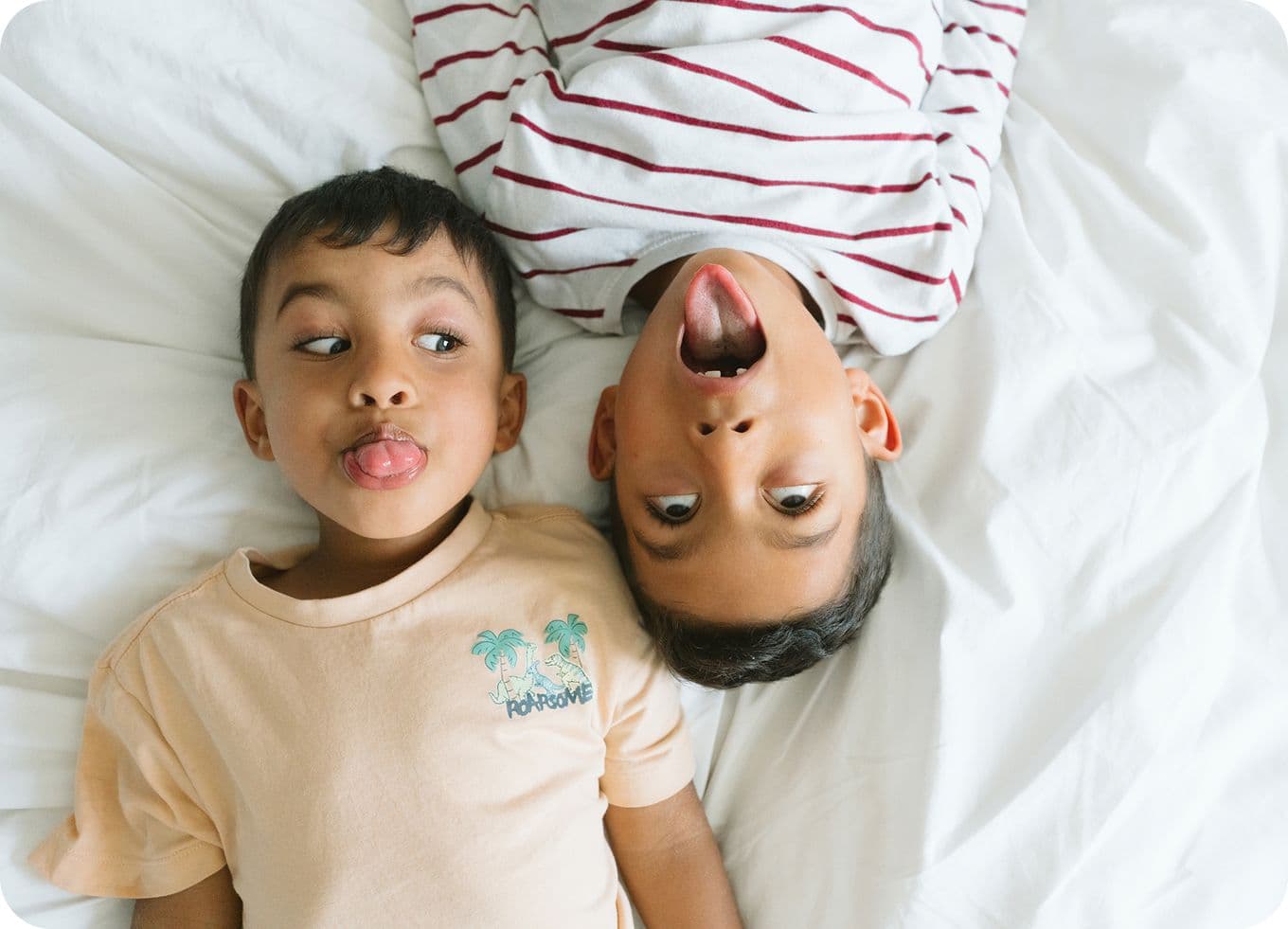 Two young boys lying on a bed, playfully sticking out their tongues, with one wearing a striped shirt and the other a palm tree t-shirt.