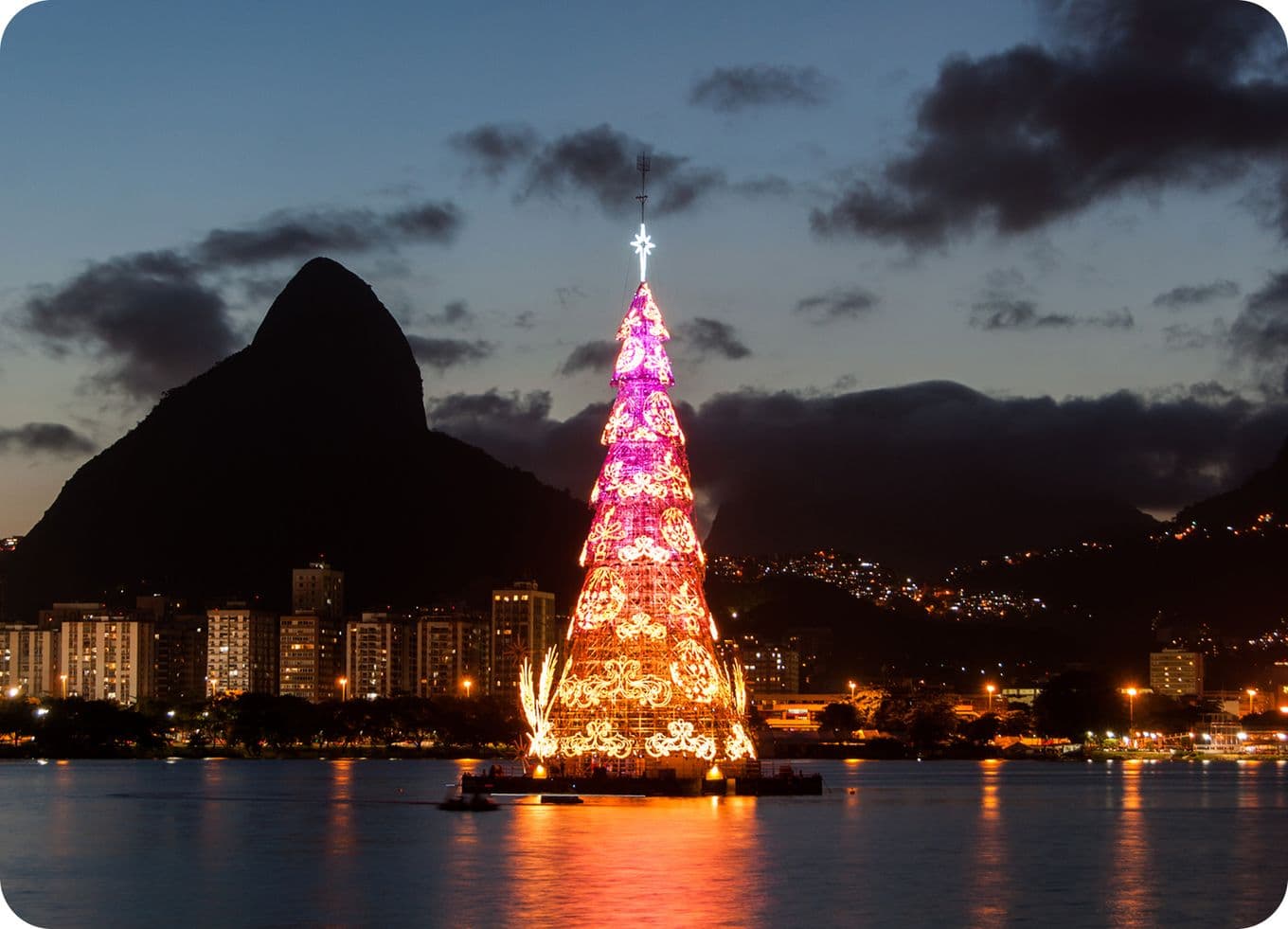 Illuminated Christmas tree on water at dusk, with city lights and a mountain silhouette in the background.
