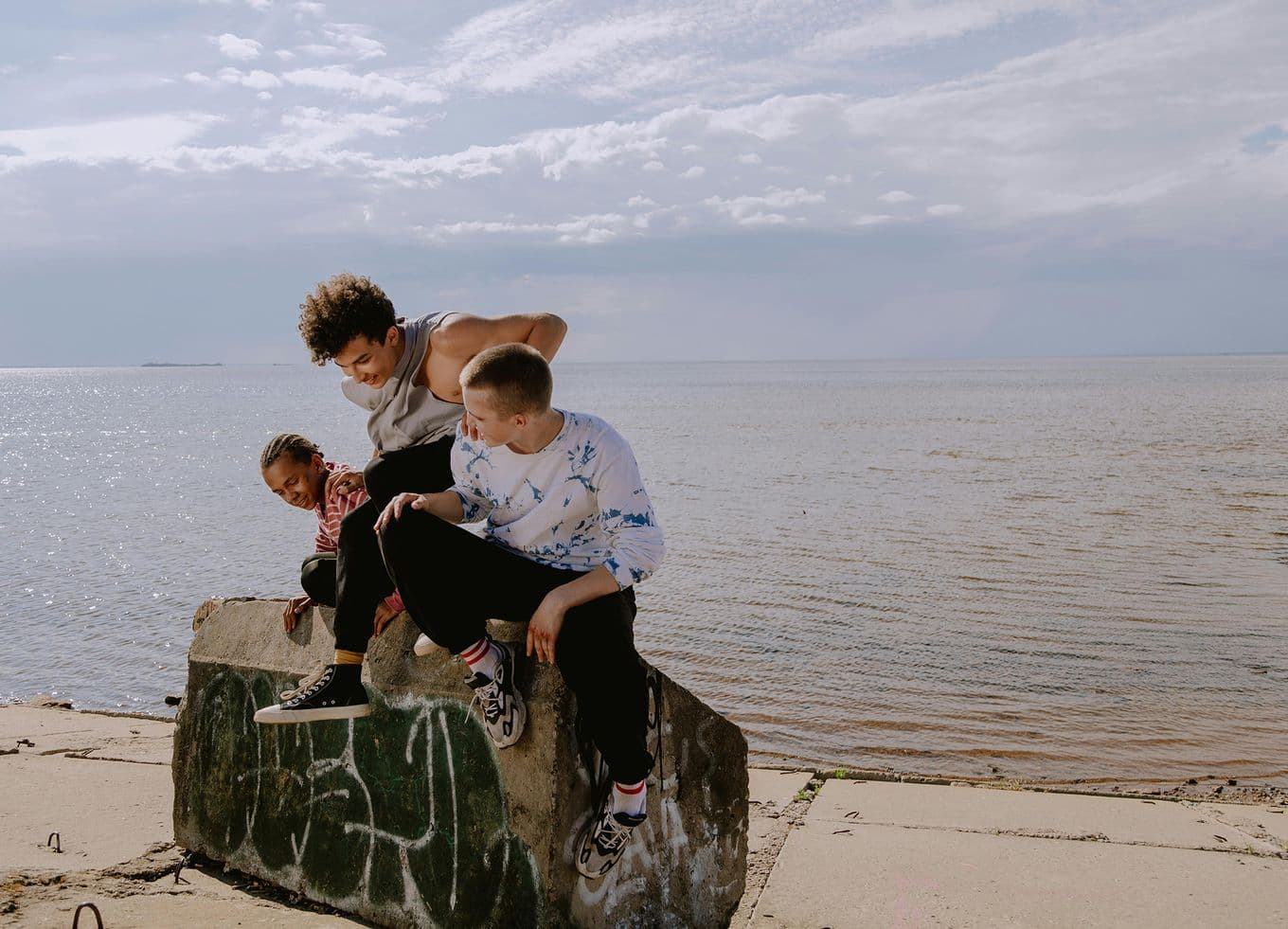 Three young people sitting on a graffiti-covered concrete block at a quiet beach, chatting and laughing under a cloudy sky.