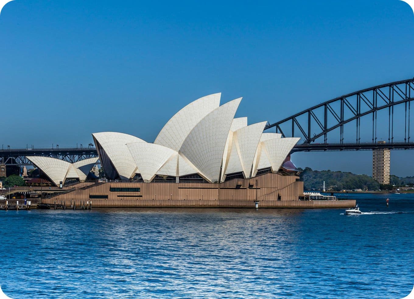 Sydney Opera House with its iconic white sails, set against a clear blue sky and water, with the Harbour Bridge in the background.