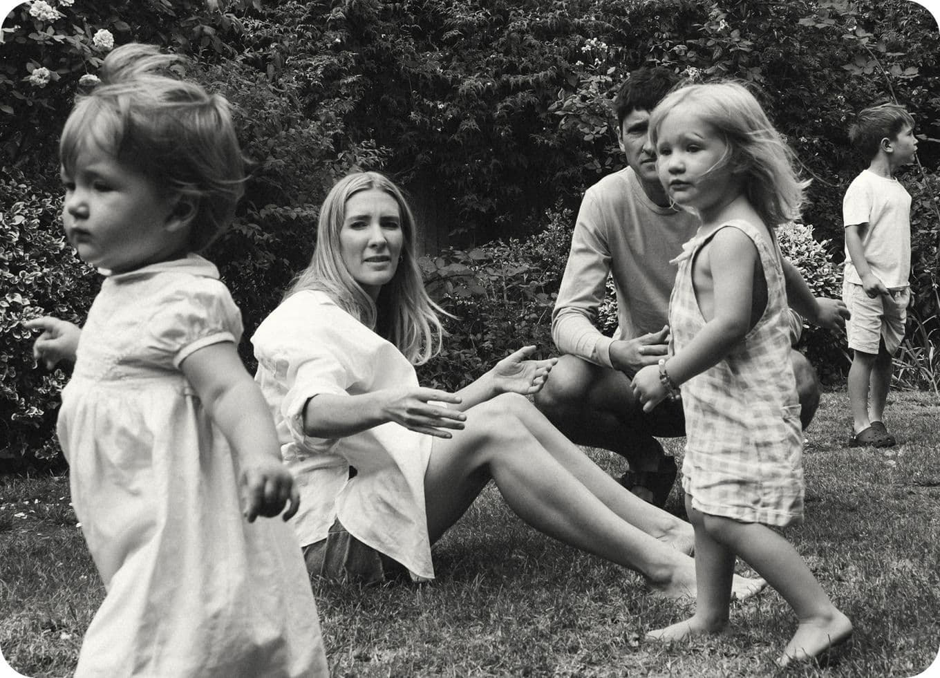 A black and white photo of a family in a garden: two adults sitting on the grass and three young children playing around them.