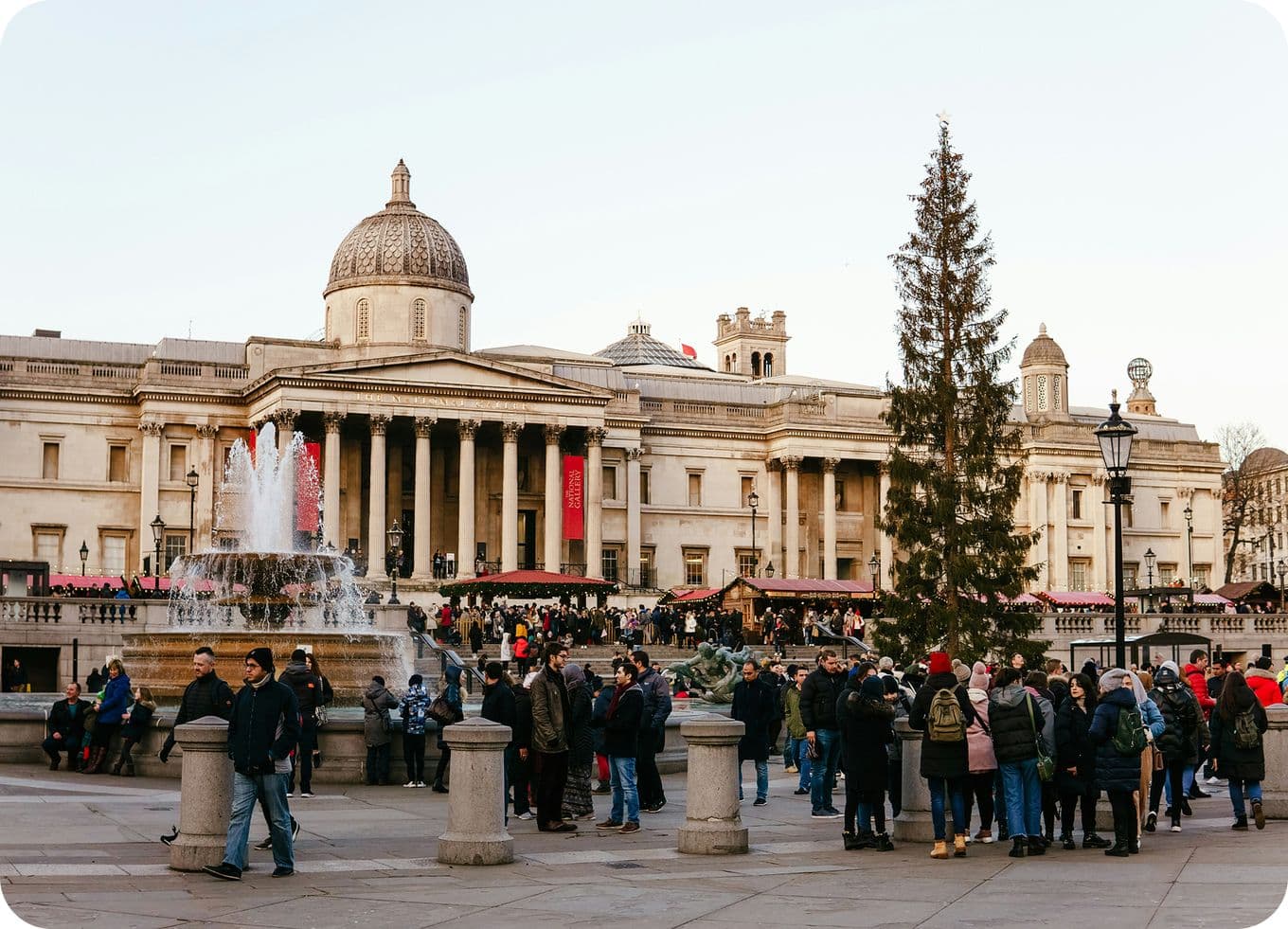 Crowd in Trafalgar Square outside the National Gallery, with fountains running and a tall Christmas tree.