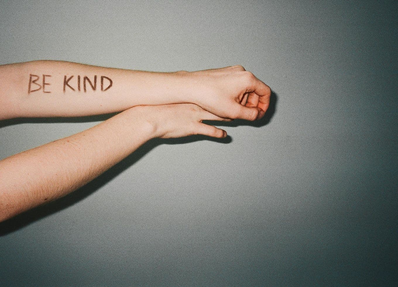 Two outstretched forearms against a gray wall, "BE KIND" written in marker on the top arm.