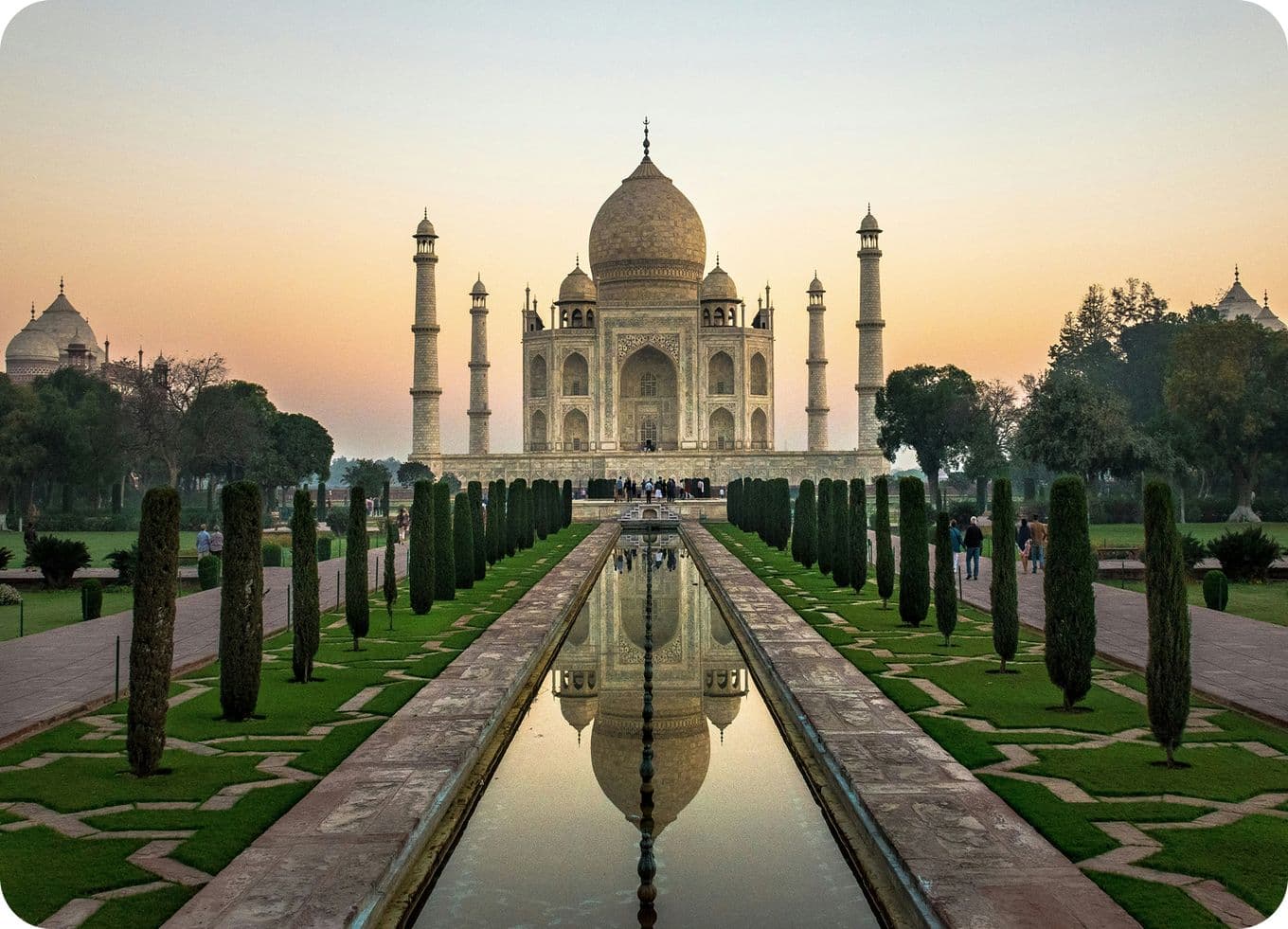 The Taj Mahal at sunrise, with its reflection in the long pool, surrounded by gardens and trees under a clear sky.