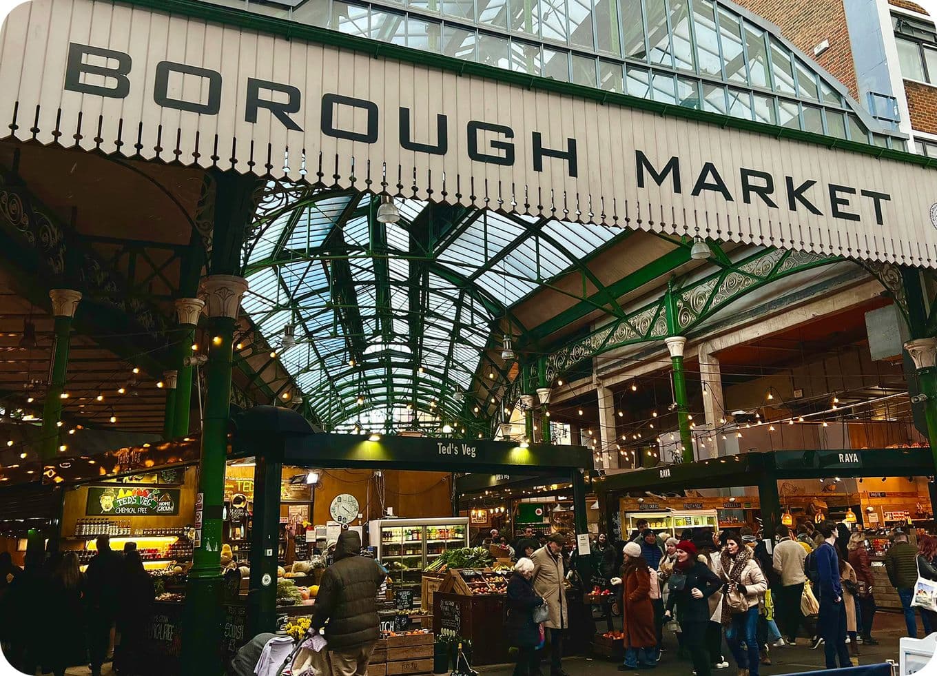 Borough Market interior under arched glass roof, green iron supports, bustling food stalls and shoppers.