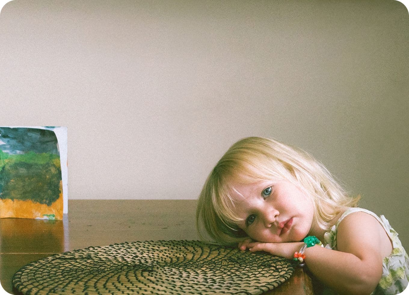 A young child with blonde hair rests their head on a table, looking at the camera. A colorful painting is in the background.