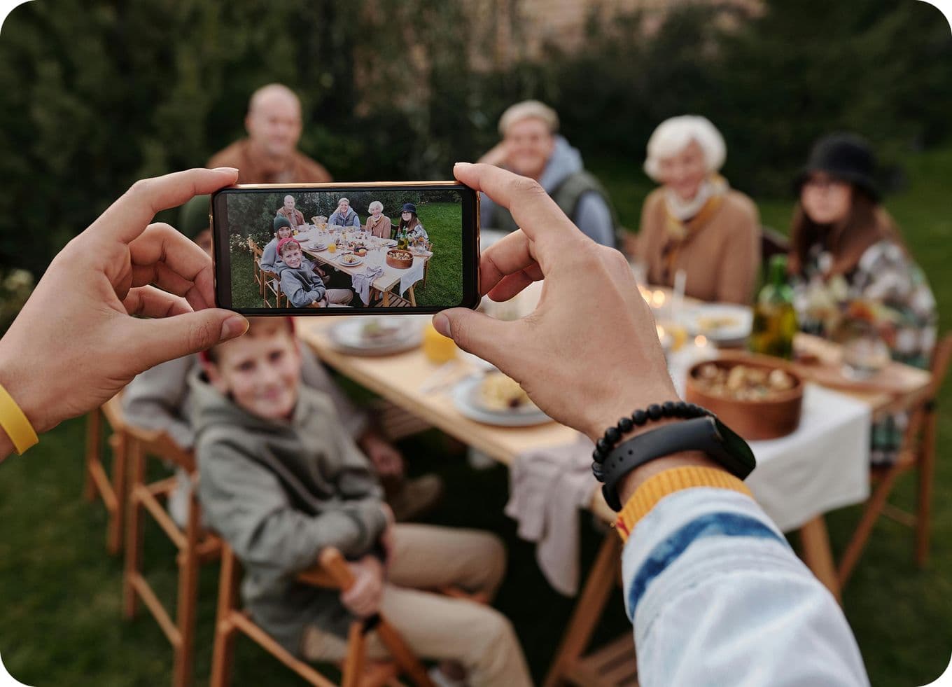 Person taking a photo of a family gathered around a table outdoors, smiling and enjoying a meal together.