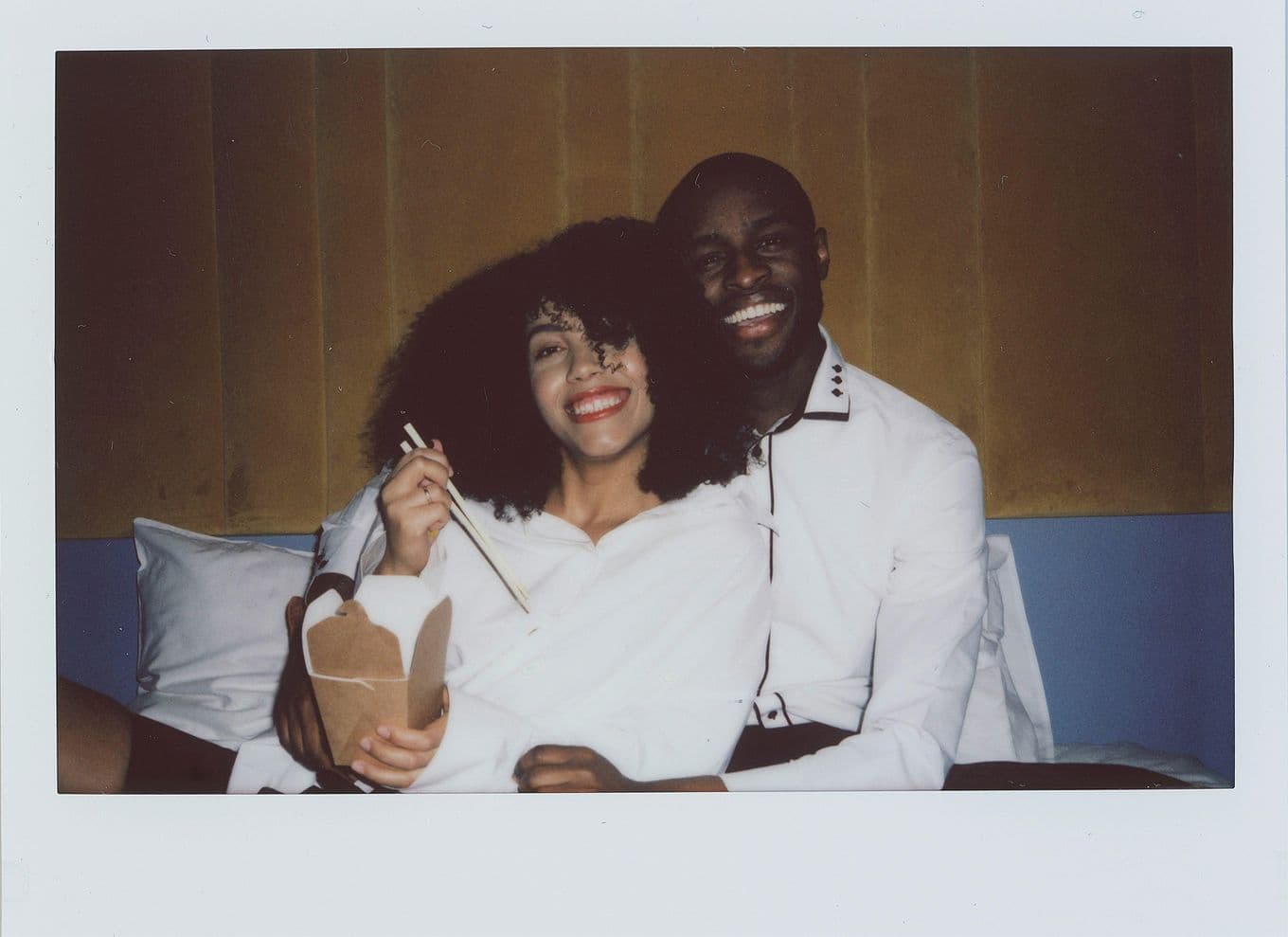 A smiling couple in white shirts sits on a bed. The woman holds chopsticks and a takeout box, while the man embraces her.