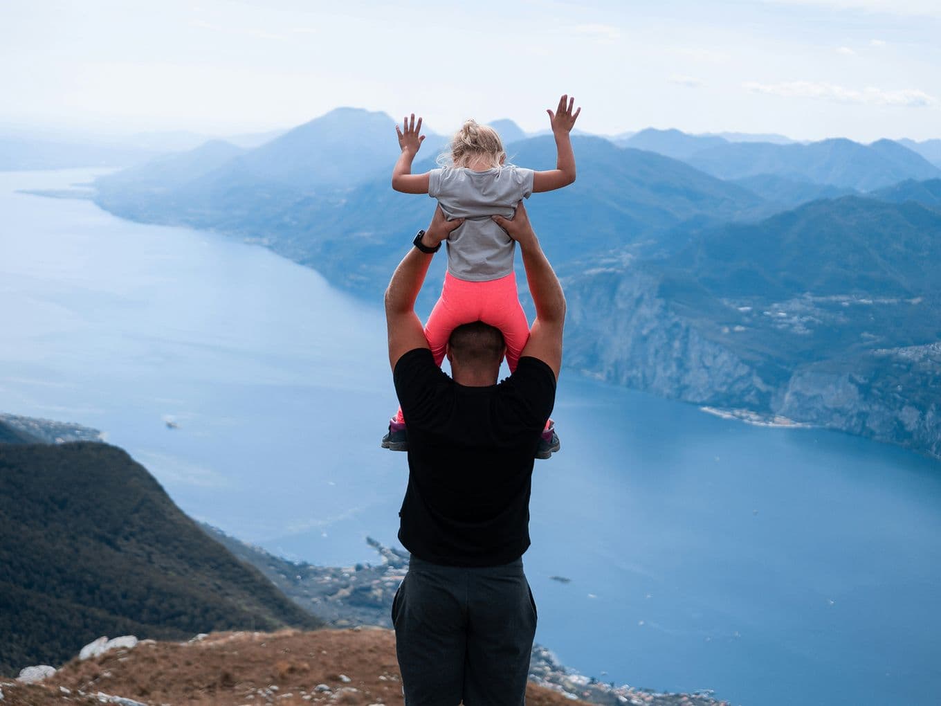Adult carrying child on shoulders, overlooking a scenic mountain and lake view. Child has arms raised, wearing pink pants and gray shirt.