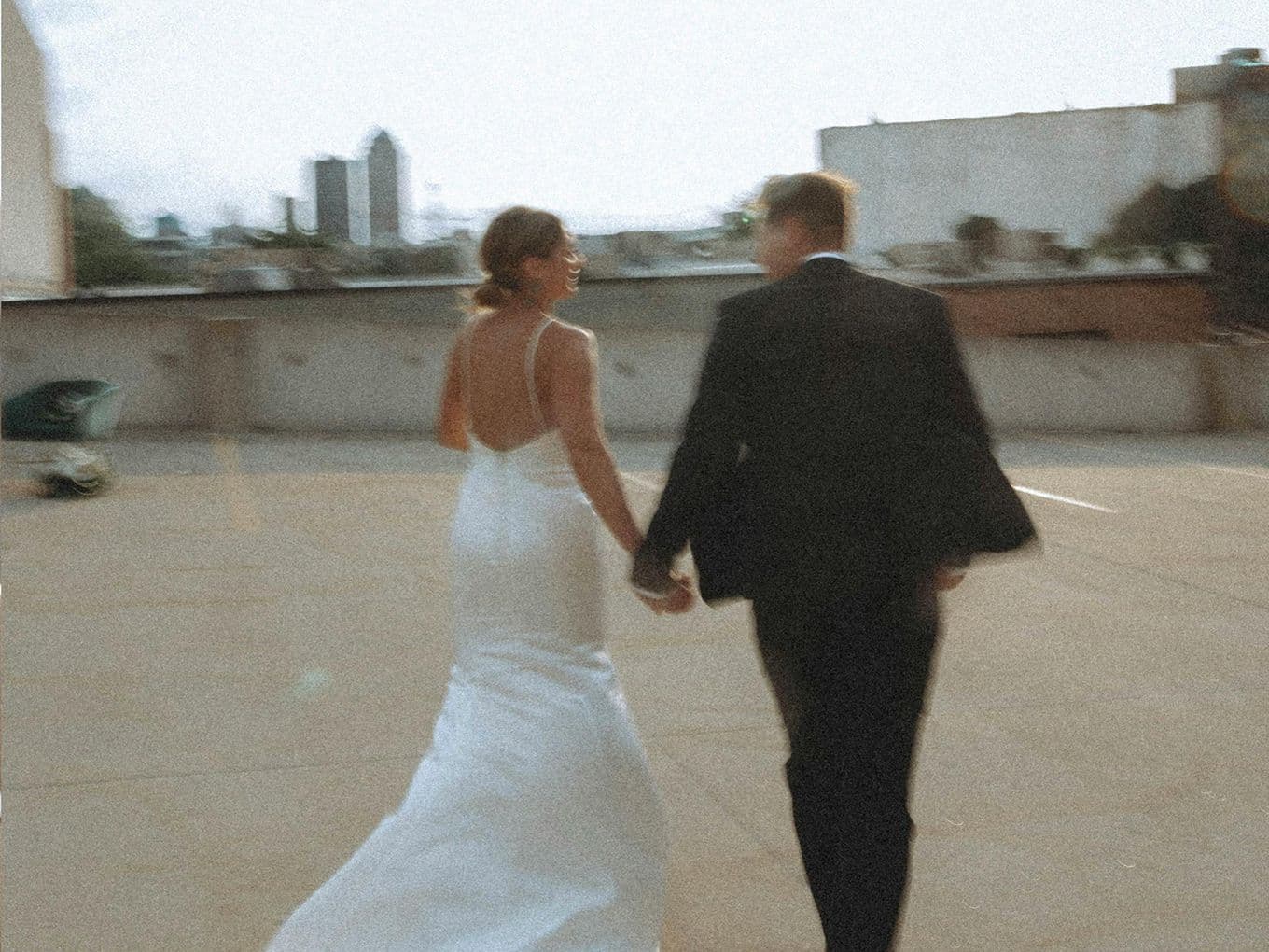 A couple in formal attire holding hands walks on a rooftop with a blurred cityscape in the background.