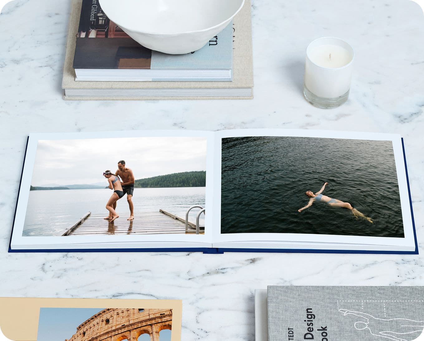 Open photo album on a marble table, showing a couple on a dock and a person swimming. Nearby are books, a candle, and a bowl.