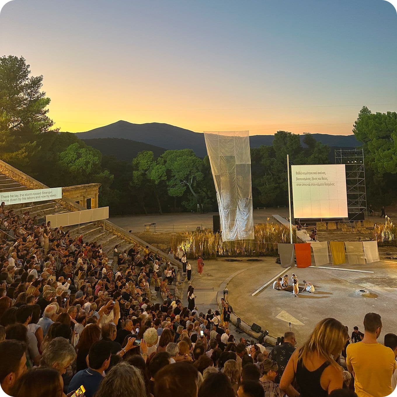 Outdoor amphitheater at sunset with a large audience seated, stage set for performance, and trees silhouetted against the sky.