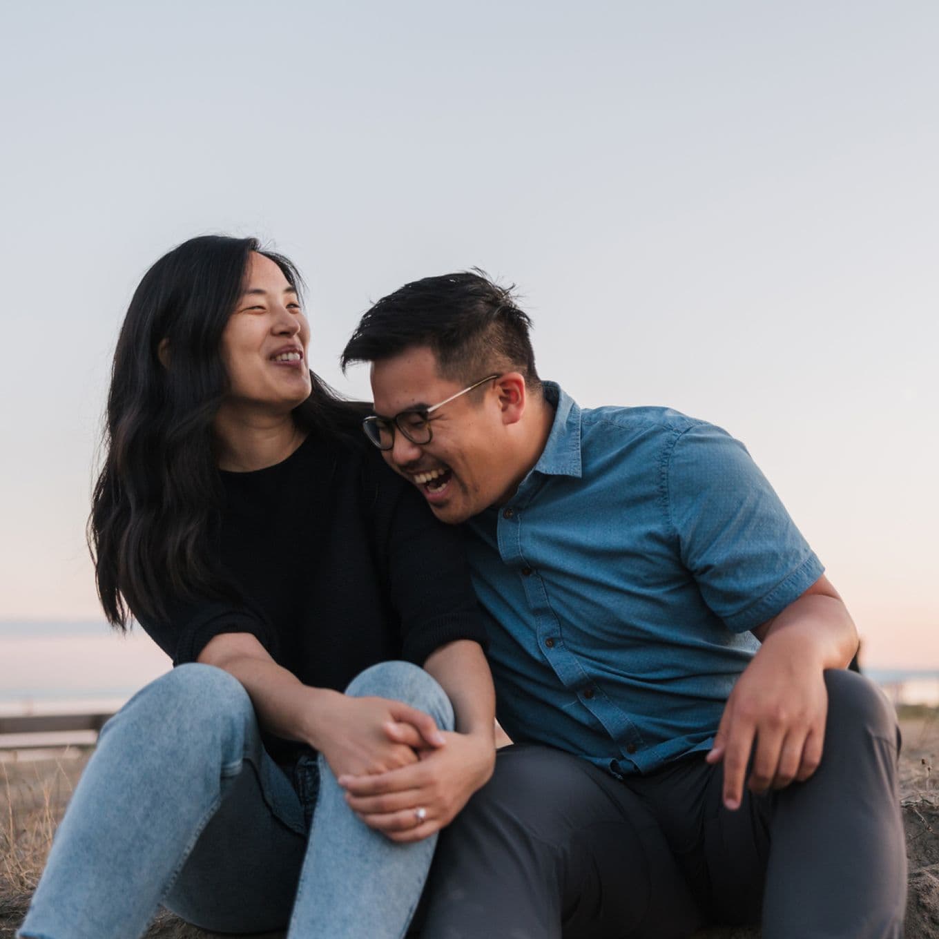 A couple sits outdoors, laughing together. The woman wears a black sweater, and the man wears a blue shirt. The sky is clear in the background.
