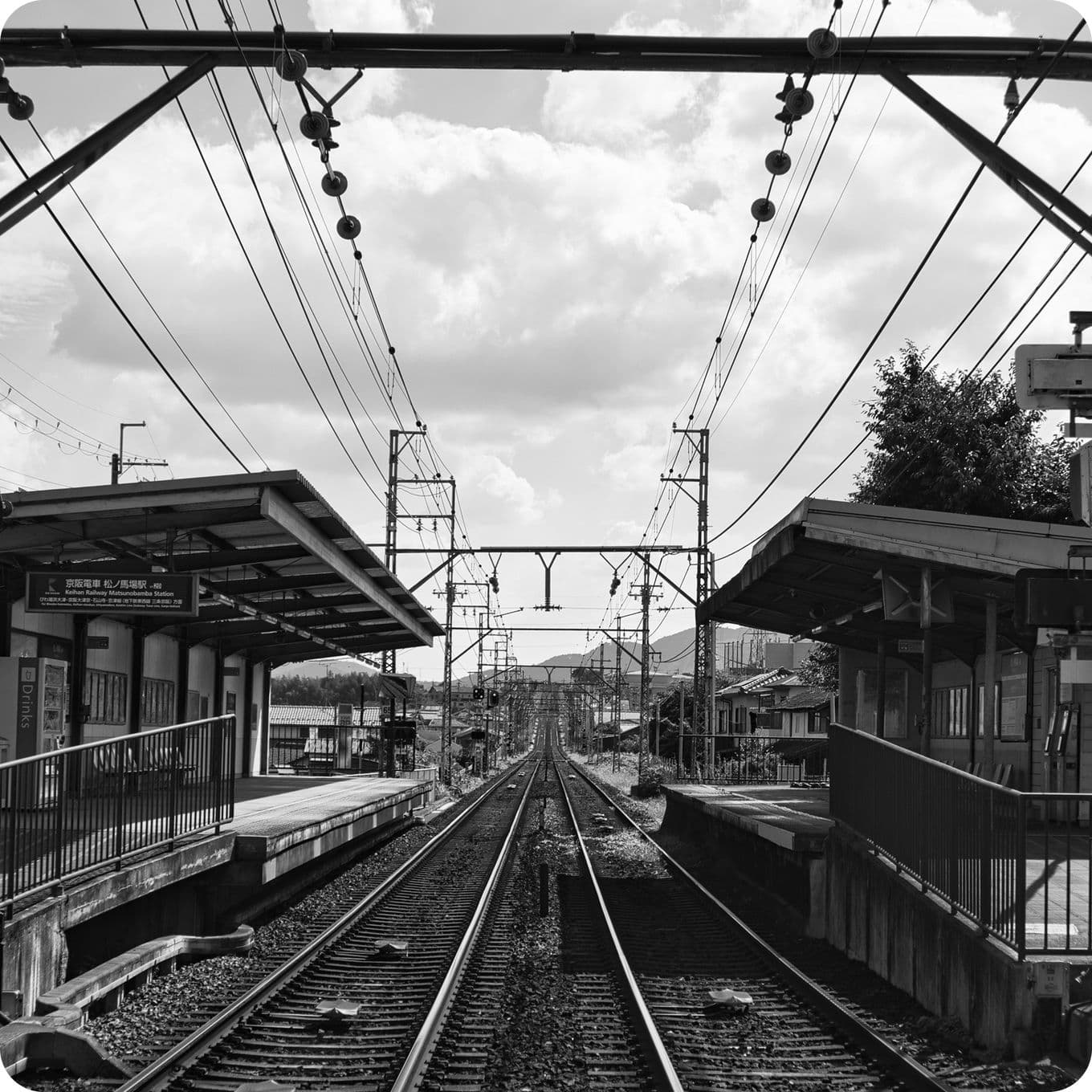 Black and white image of a railway station with parallel tracks, overhead cables, and platforms on both sides under a cloudy sky.
