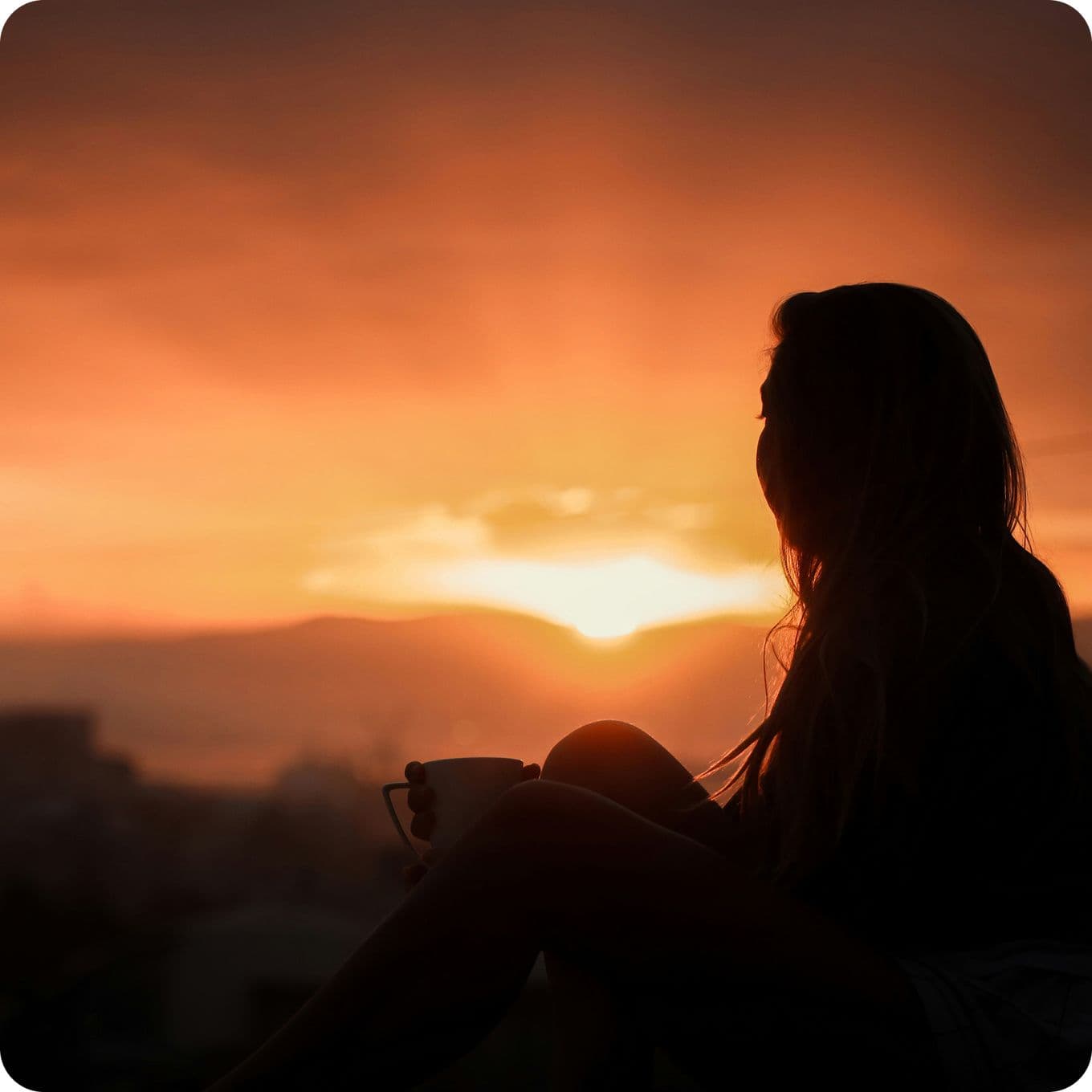 Silhouette of a person sitting with a cup, gazing at a vibrant orange sunset over distant hills.