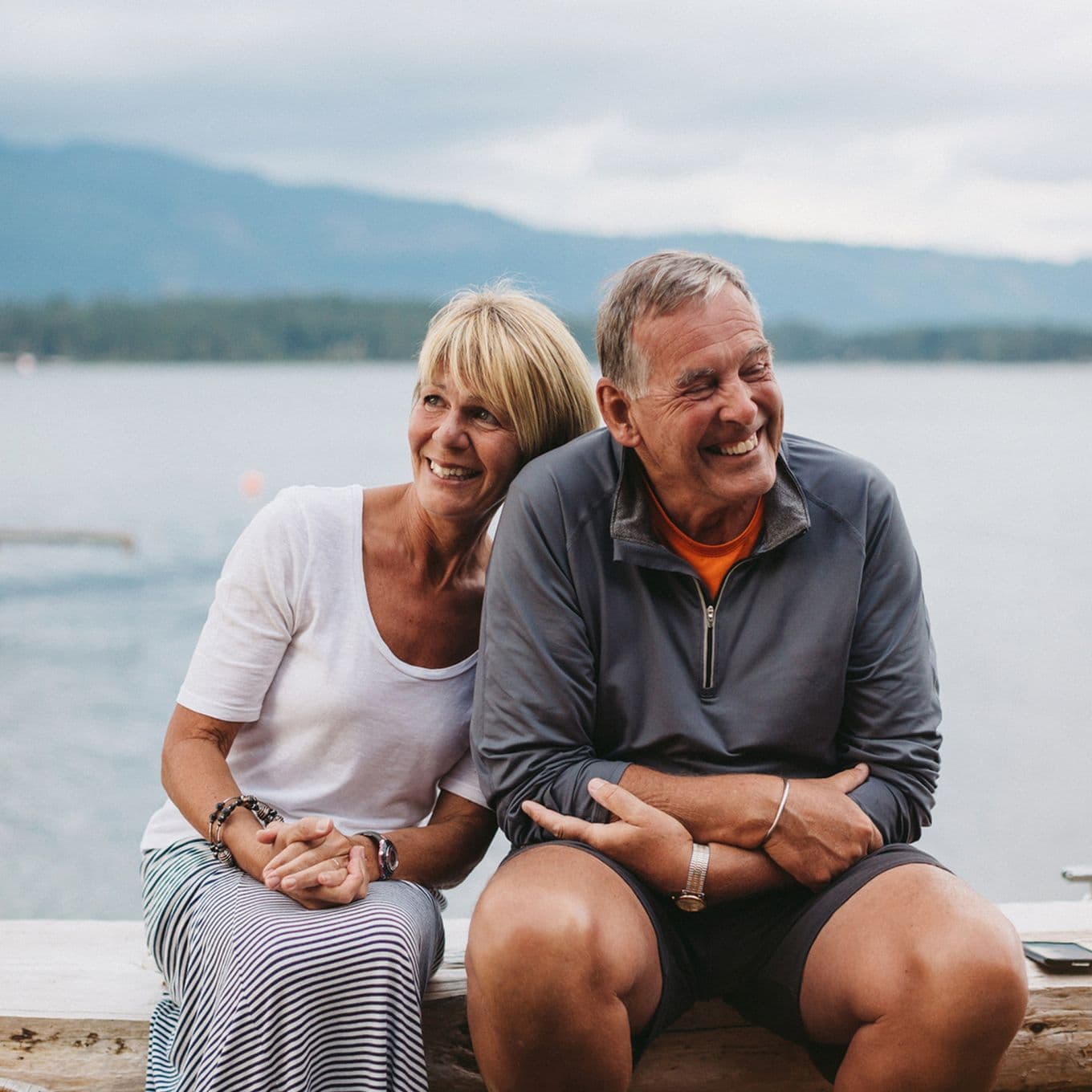 Smiling older couple sitting together by a lake, with mountains in the background. The woman leans her head on the man's shoulder.