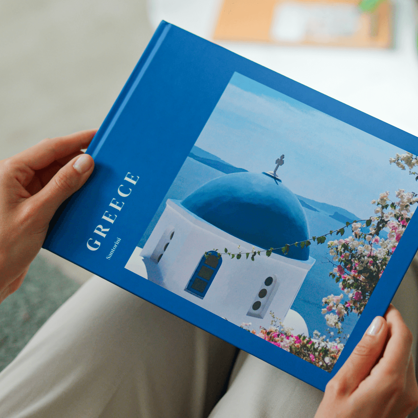 A person holding a Popsa Photo Book titled "Greece, with a blue-domed building and flowers against a sea view on the cover