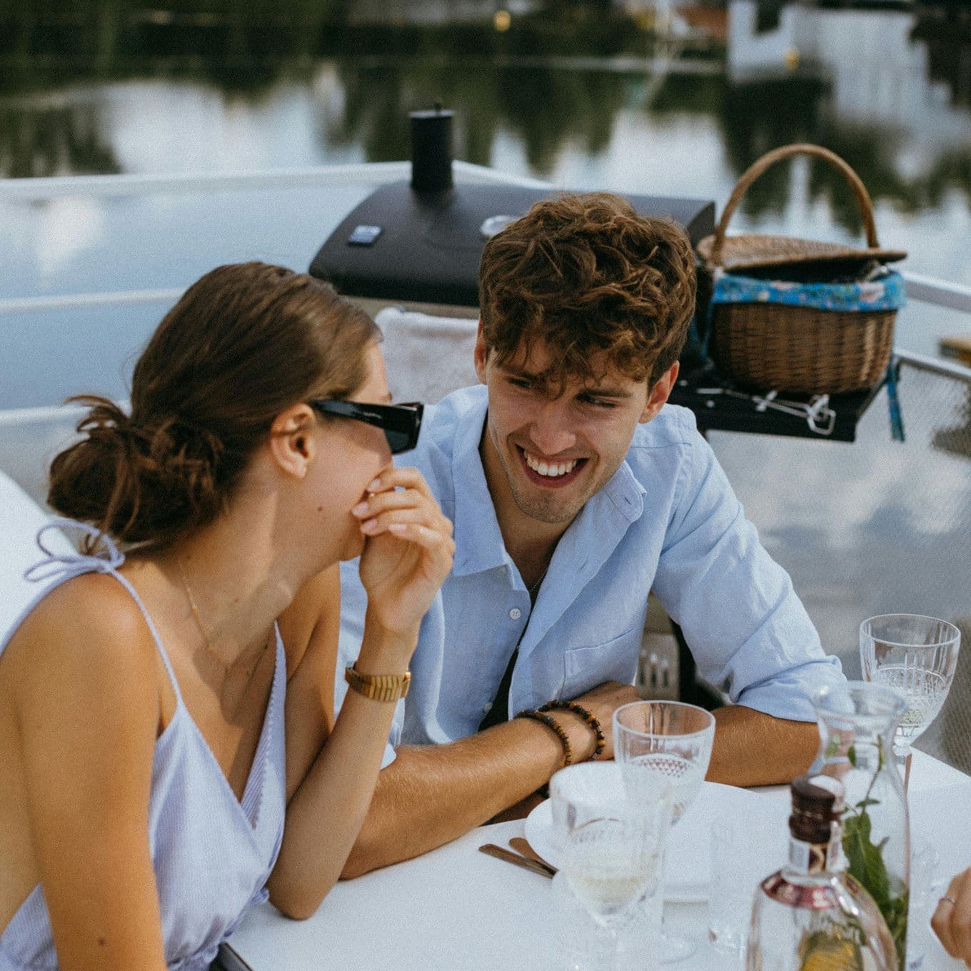 A couple in light clothing enjoys a meal on a boat, smiling at each other. The table has glasses and a bottle, with a wicker basket in the background.