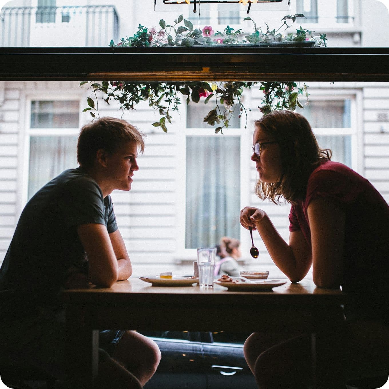 Two people sit across from each other at a cafe table, engaged in conversation, with a window and hanging plants in the background.