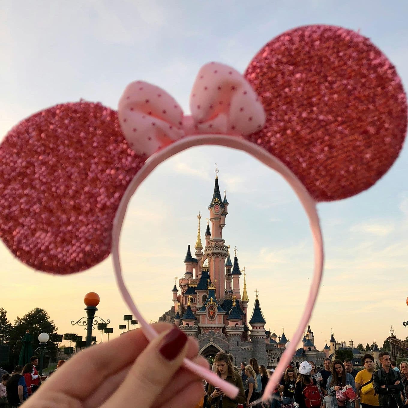 A hand holding a Minnie Mouse headband with novelty ears in front of Sleeping Beauty's Castle, Disneyland Paris, France