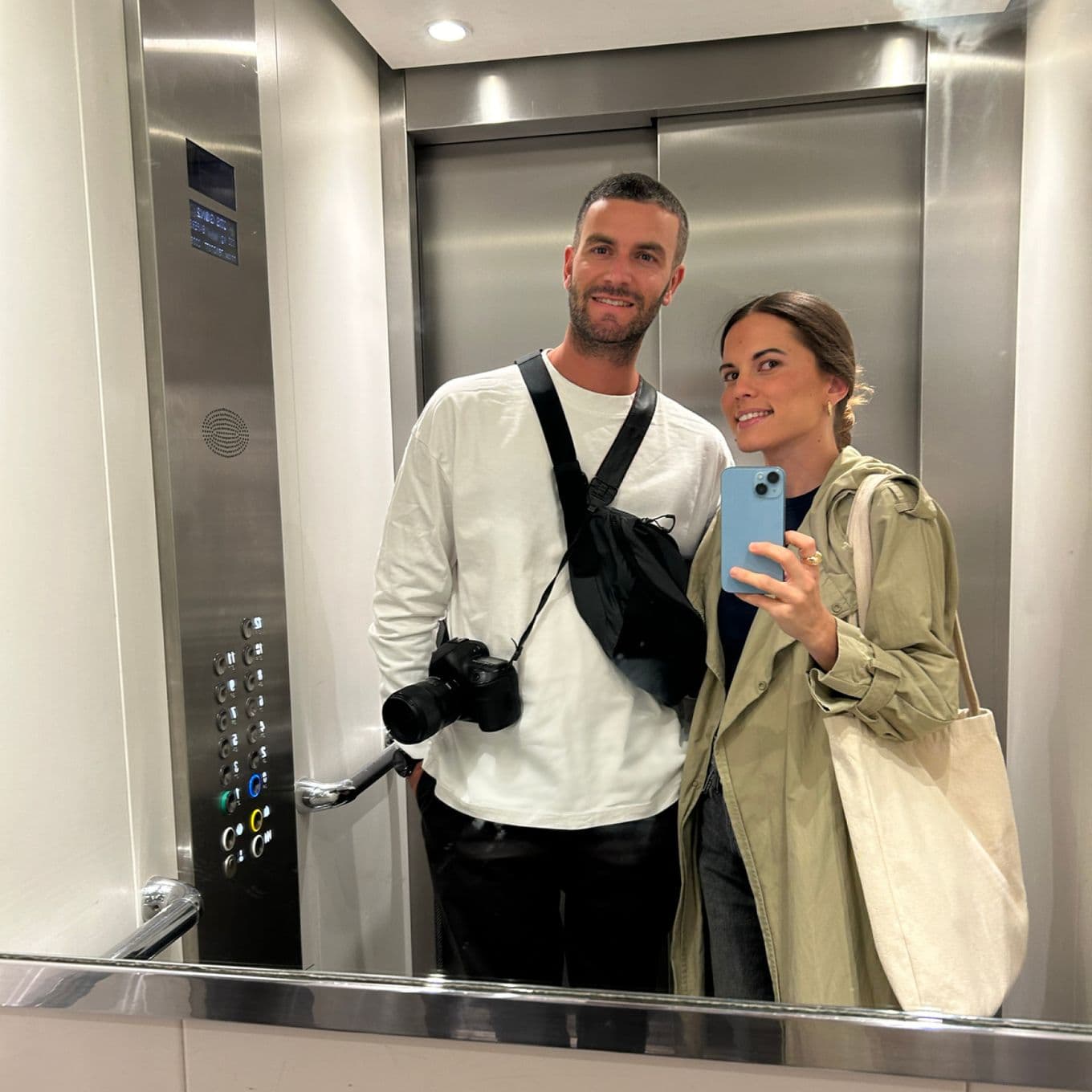 A man and woman taking a selfie in an elevator, both smiling. The man has a camera, and the woman holds a phone.