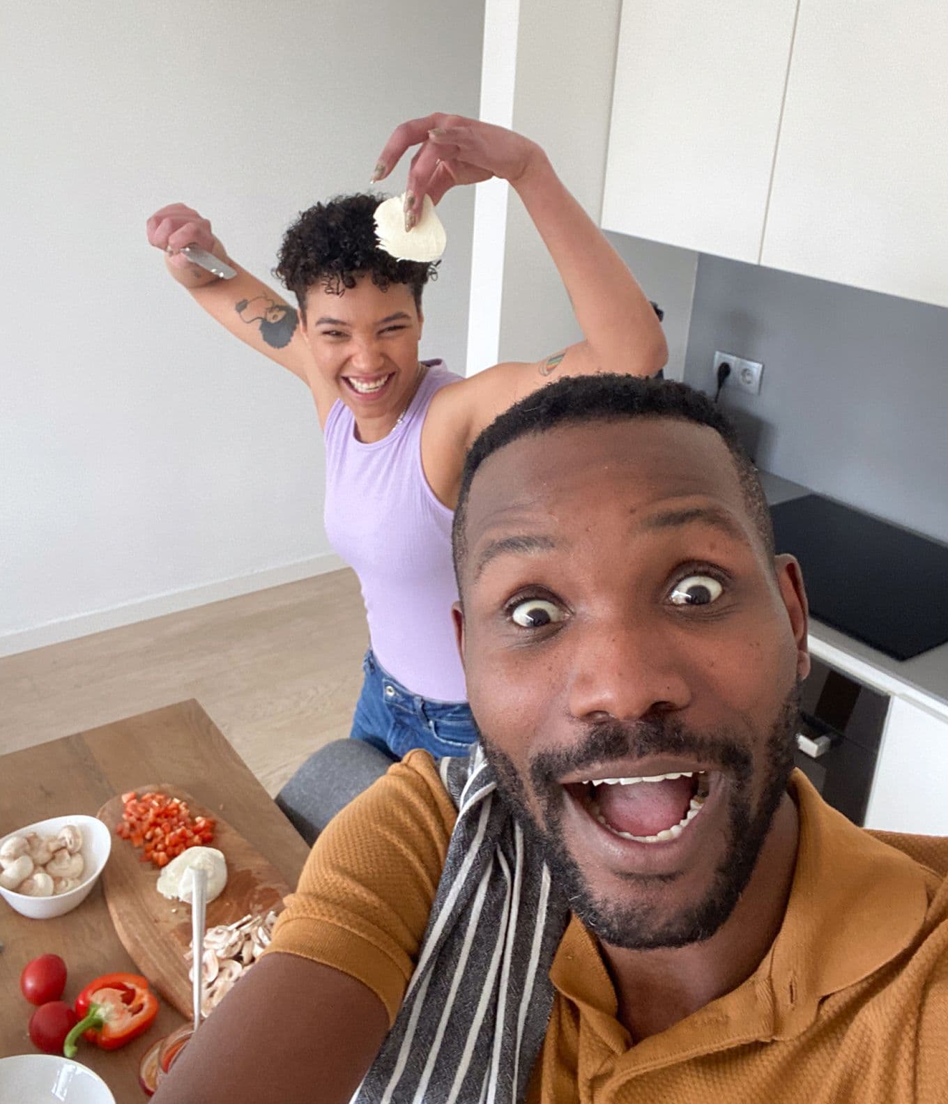 A joyful couple in a kitchen, one holding cheese and a knife playfully, with chopped vegetables on a wooden board nearby.