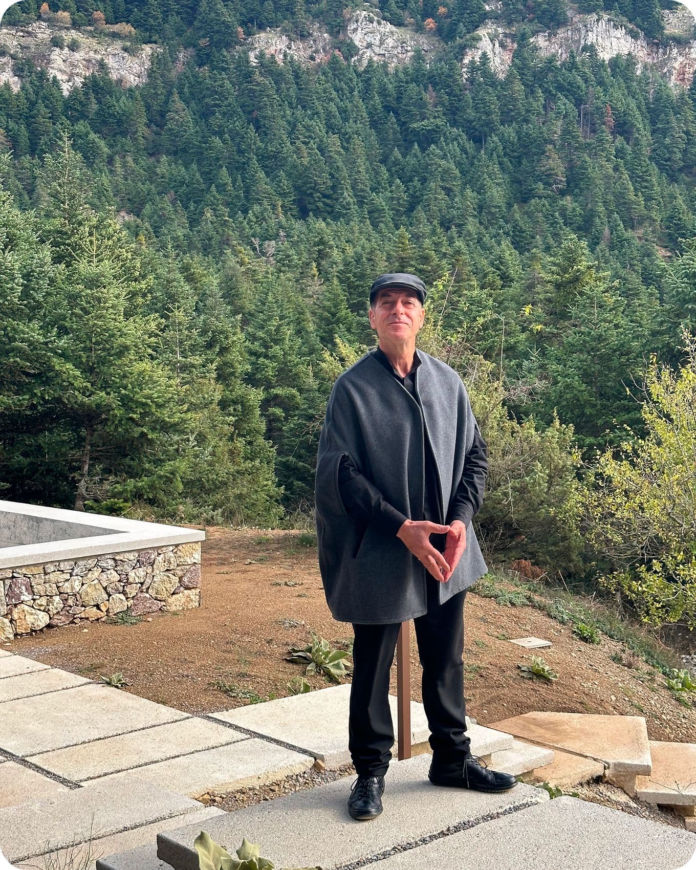 A person in a gray cape and black cap stands outdoors on a stone path, with a forested mountain landscape in the background.