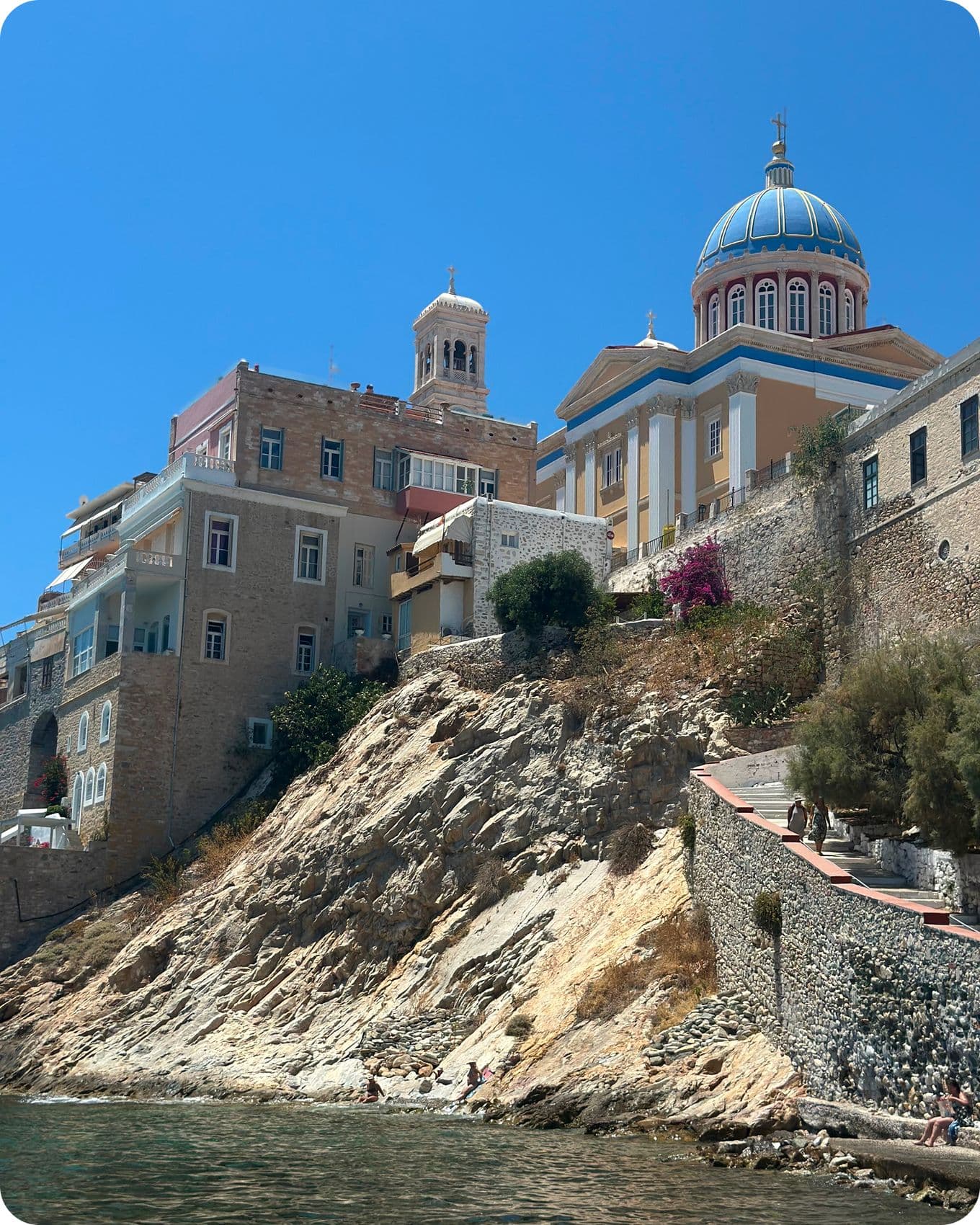 View of colorful buildings and a domed church perched on a rocky hillside by the sea under a clear blue sky.