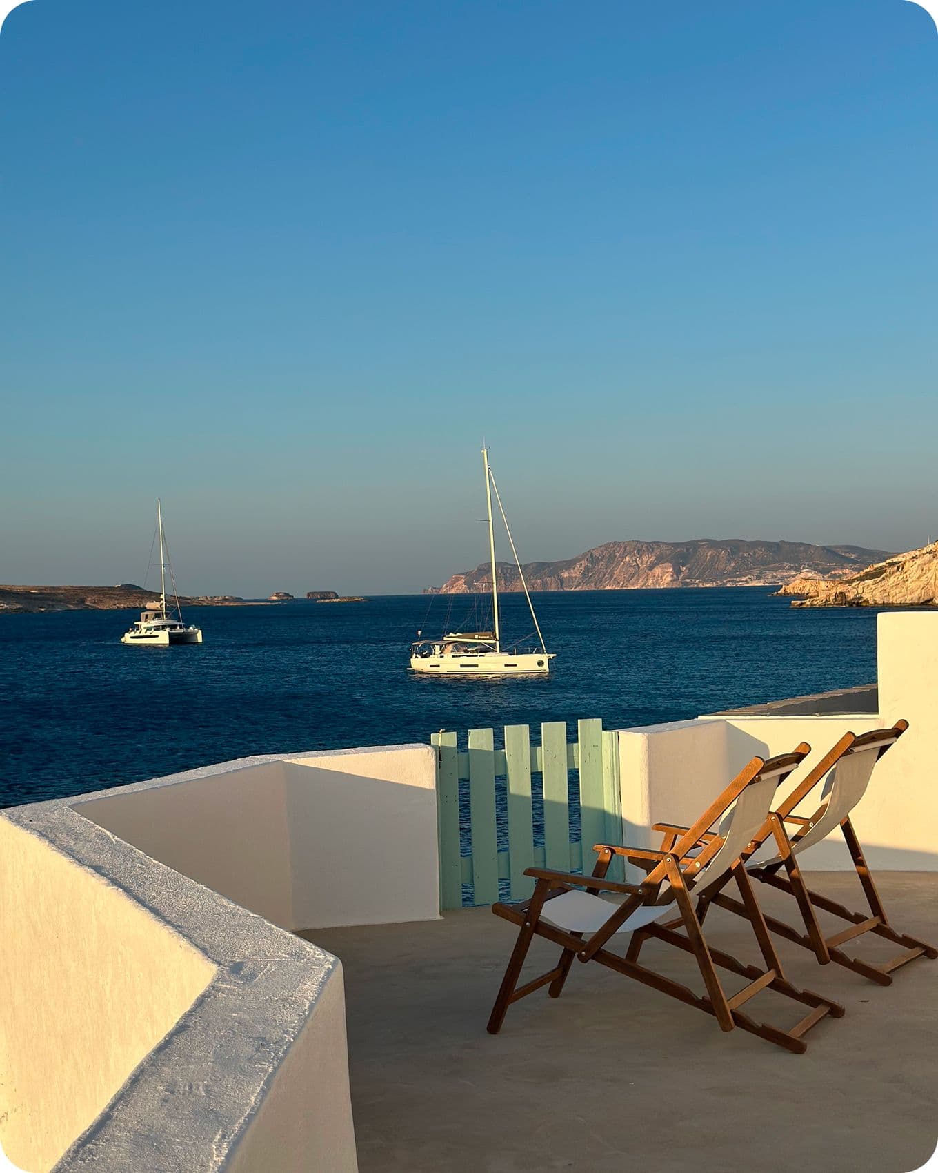 Two wooden chairs on a white terrace overlook a tranquil sea with sailboats and a distant rocky coastline under a clear blue sky.
