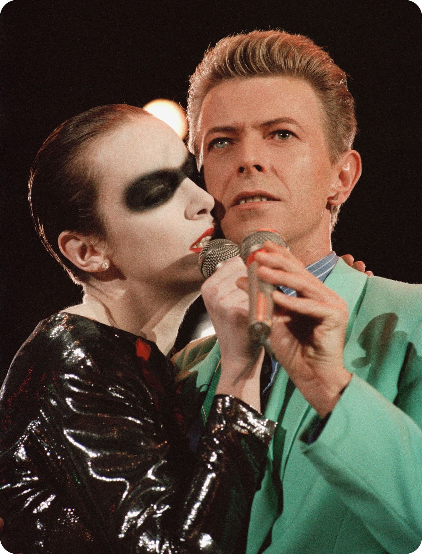 Two performers on stage, Annie Lennox with dramatic makeup and black outfit, and David Bowie in a turquoise suit, both holding microphones closely.