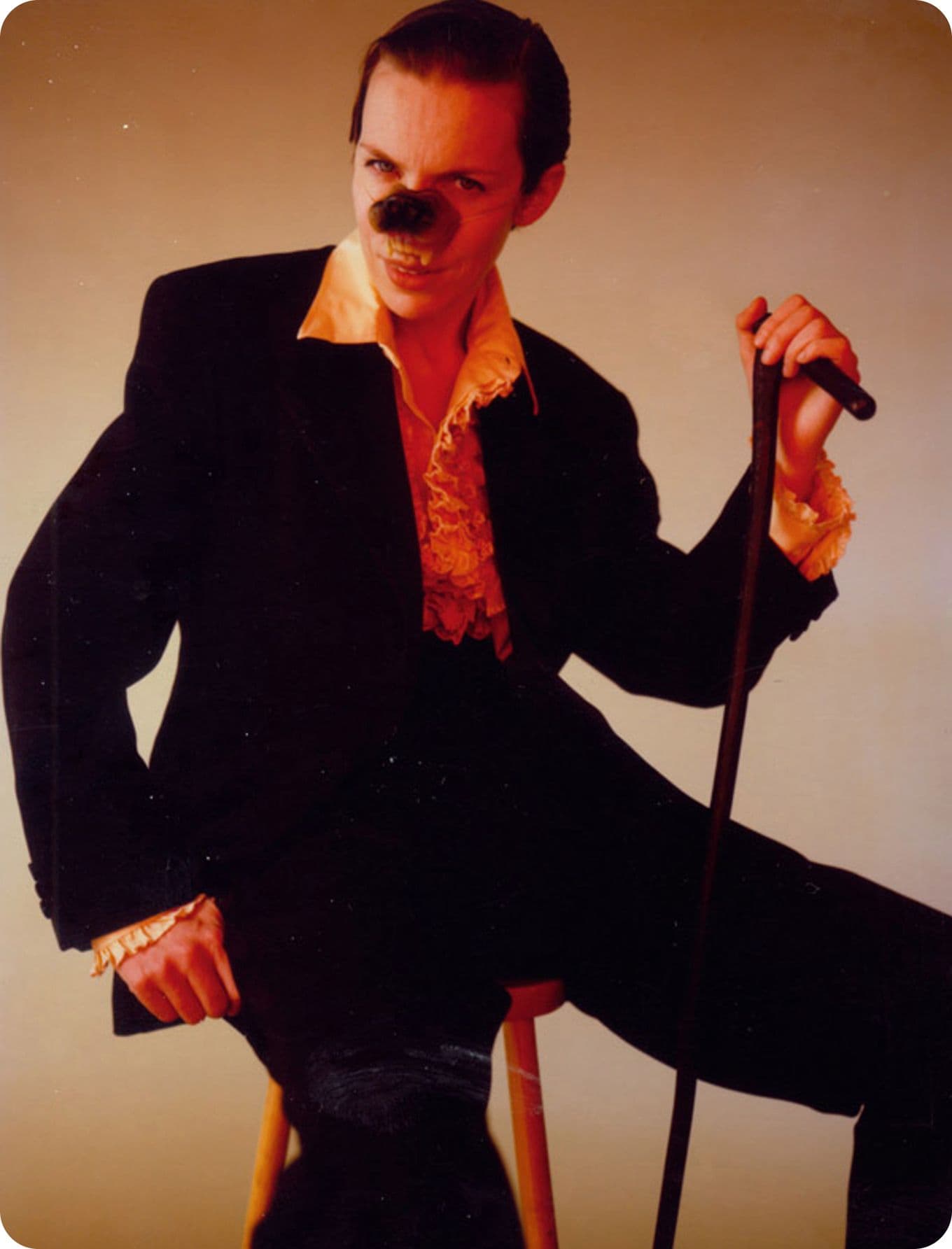 Annie Lennox in a dark suit with a ruffled shirt, sitting on a stool, holding a cane, and wearing a fake nose in a dramatic pose.