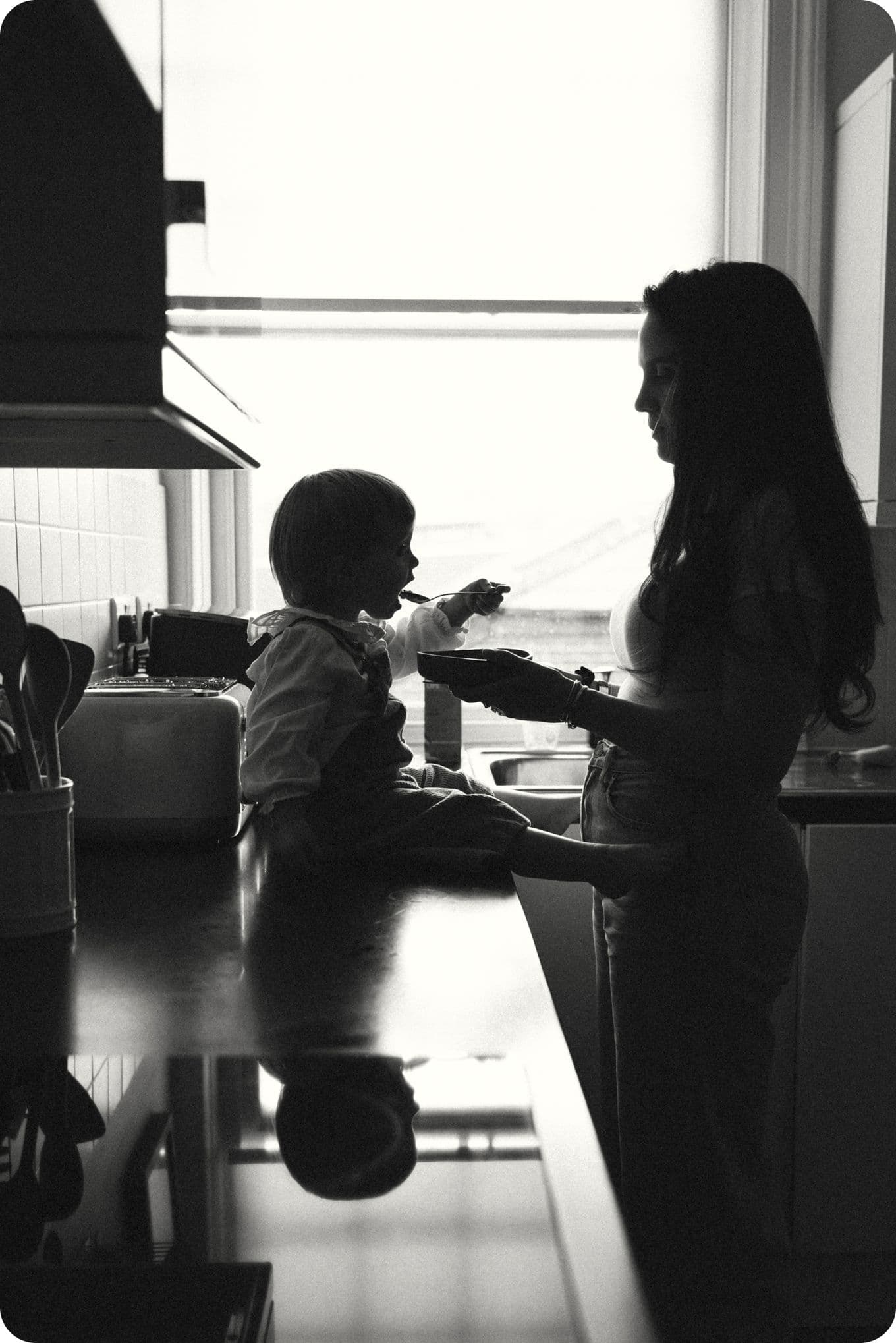 Silhouette of a woman feeding a child sitting on a kitchen counter, with light streaming through a window in the background.