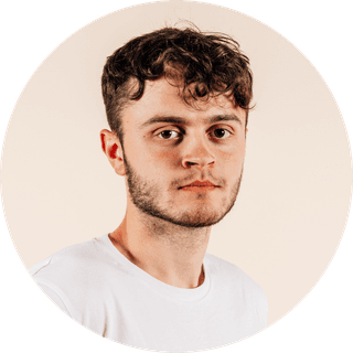 Young man with short curly hair and a beard, wearing a white t-shirt, against a neutral background, looking directly at the camera.