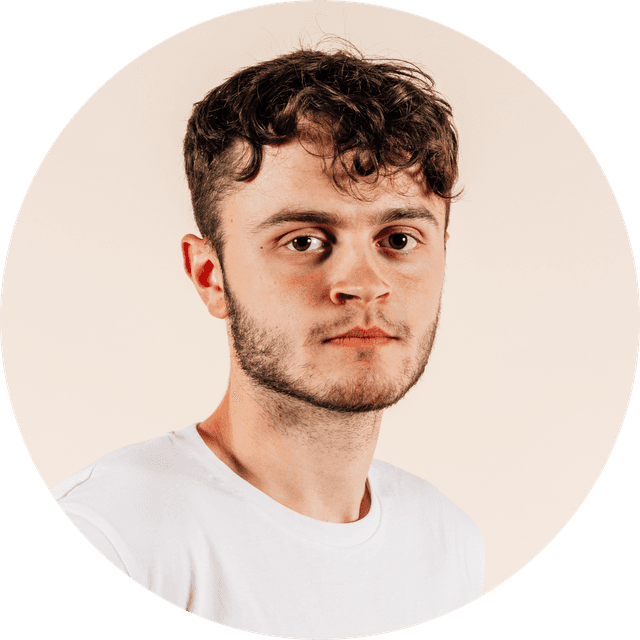 Young man with short curly hair and a beard, wearing a white t-shirt, against a neutral background, looking directly at the camera.