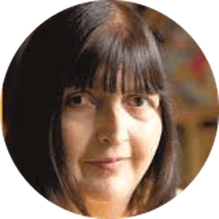 A woman with straight, dark hair and bangs, looking at the camera with a neutral expression. Blurred background.