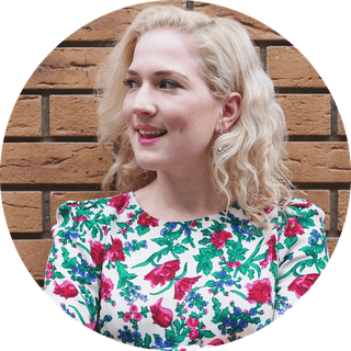 Young woman with wavy blonde hair smiling, wearing a white floral dress, looking left against a brick wall.