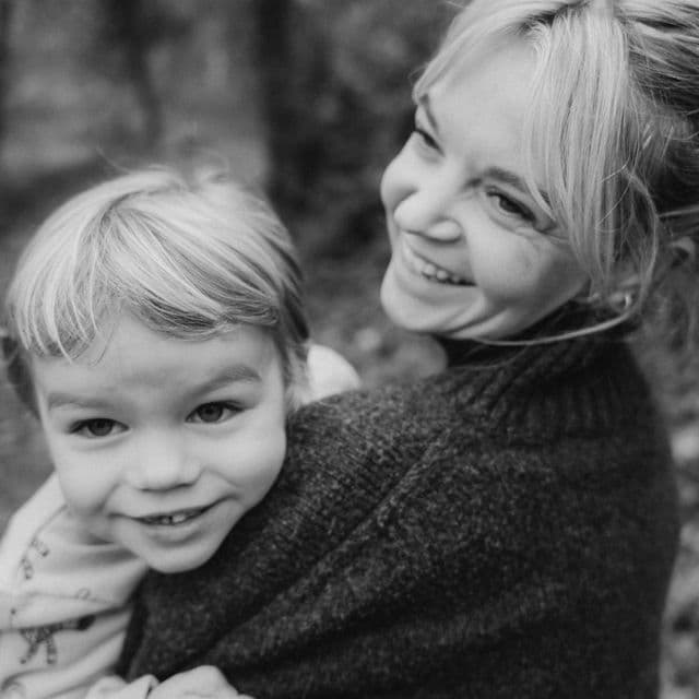 A woman smiling while carrying a young child on her back in a forest. Black and white photo.