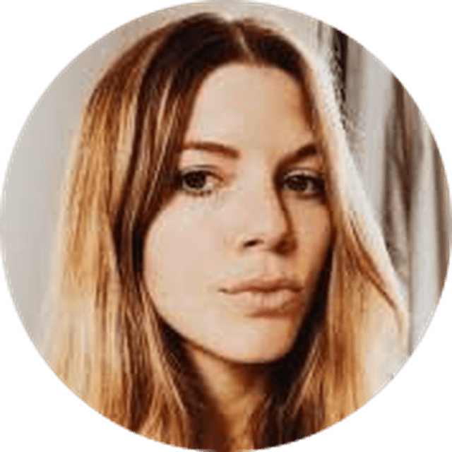 A woman with long, light brown hair and a neutral expression looking at the camera. She is indoors with soft lighting.