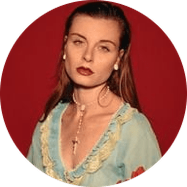Woman with long hair, wearing a light blue dress with lace trim and a necklace, stands against a red background.