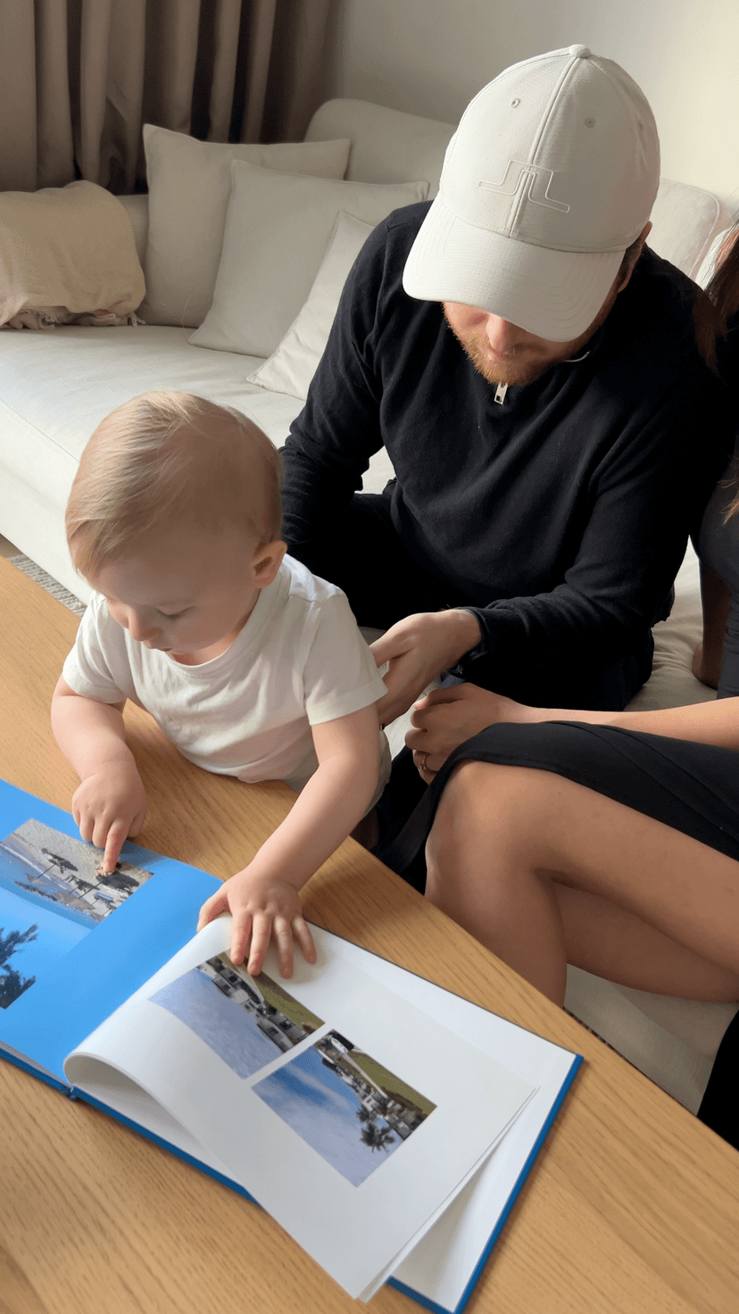 A toddler points at a photo in a photo book on a table, sitting with two adults. The scene is cozy, with a sofa and cushions in the background.