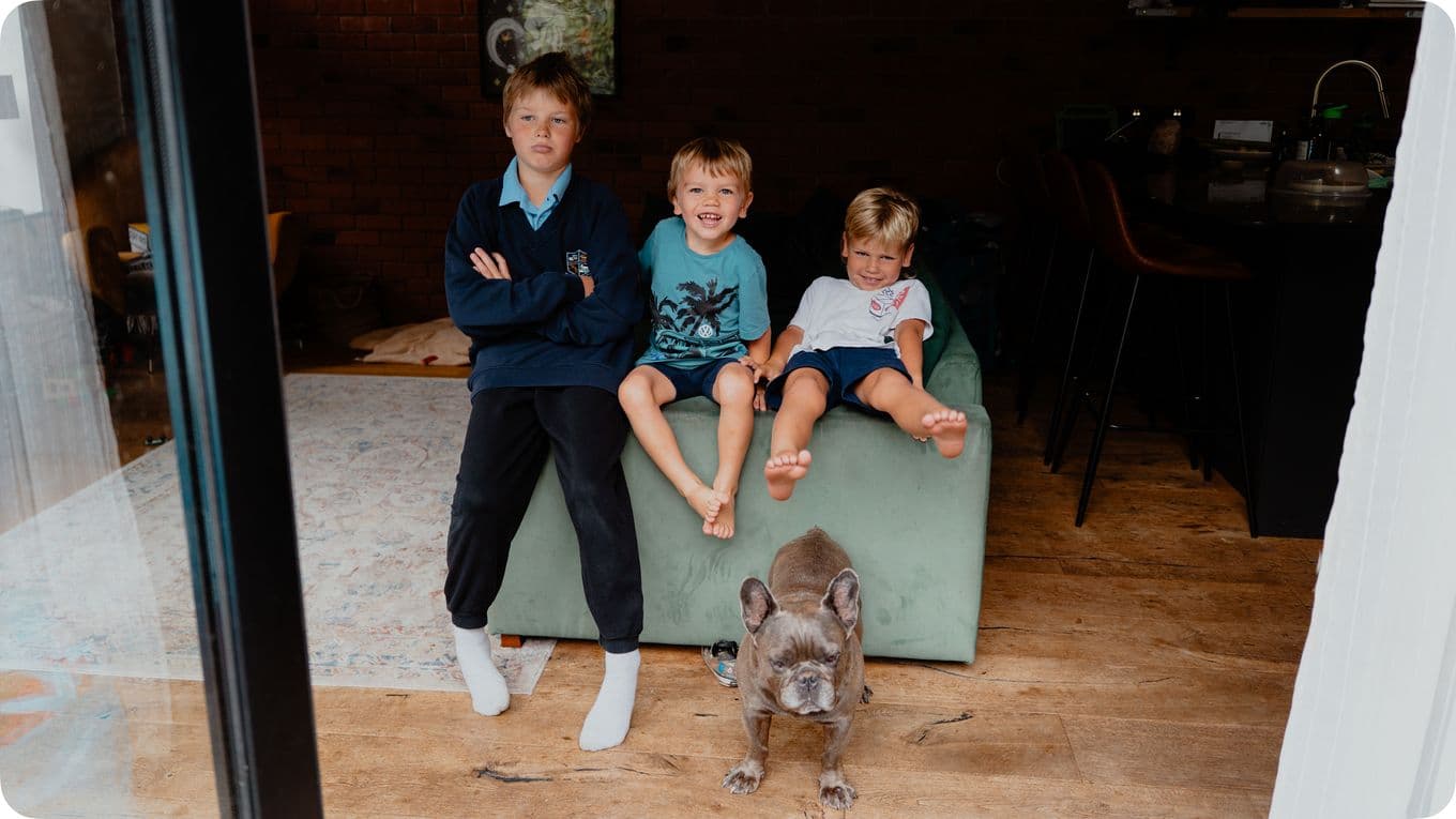 Three young boys sit on a couch, smiling, with a gray dog standing on the wooden floor in front of them.