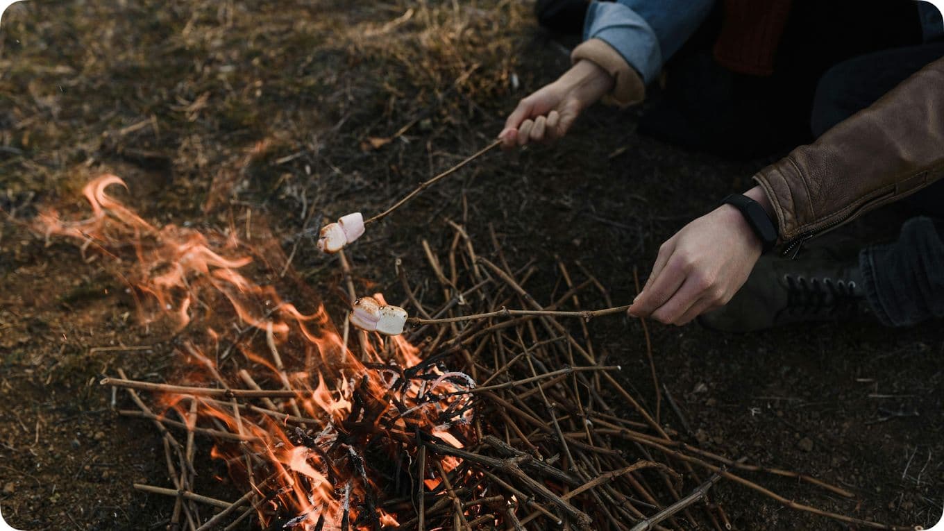 Two people roasting marshmallows over a small campfire with sticks, surrounded by twigs and dry grass.