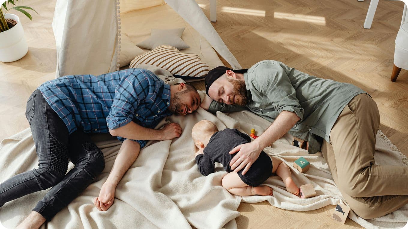 Two adults and a baby lie on a blanket on the floor, surrounded by toys, under a tent in a cozy room.