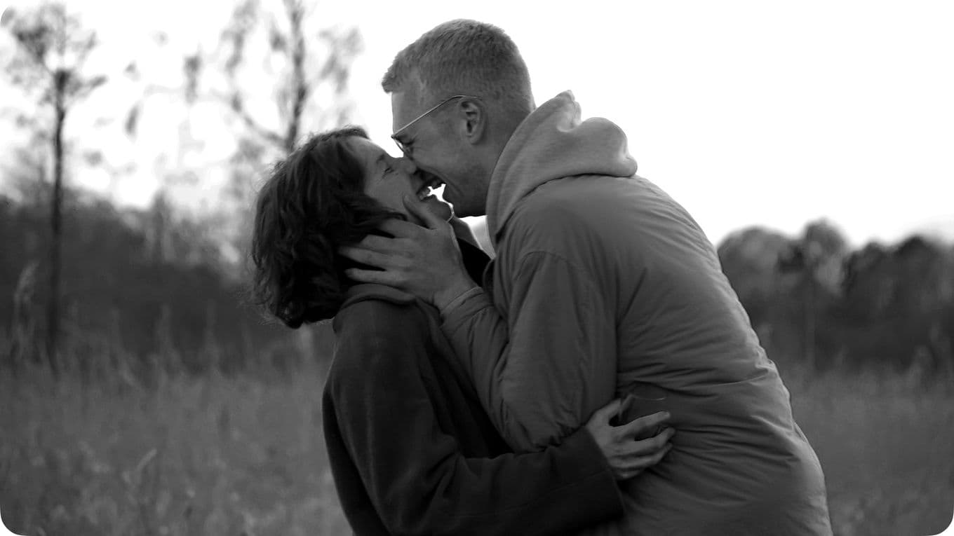 A couple embraces and smiles at each other in a field, surrounded by tall grass and trees, in a black and white photo.