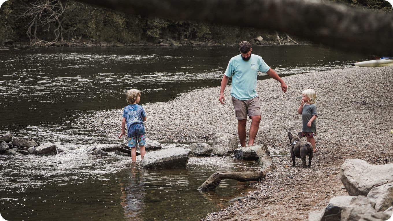 A man and two children walk along a rocky riverbank with a dog. One child wades in the water while the others stay on the shore.